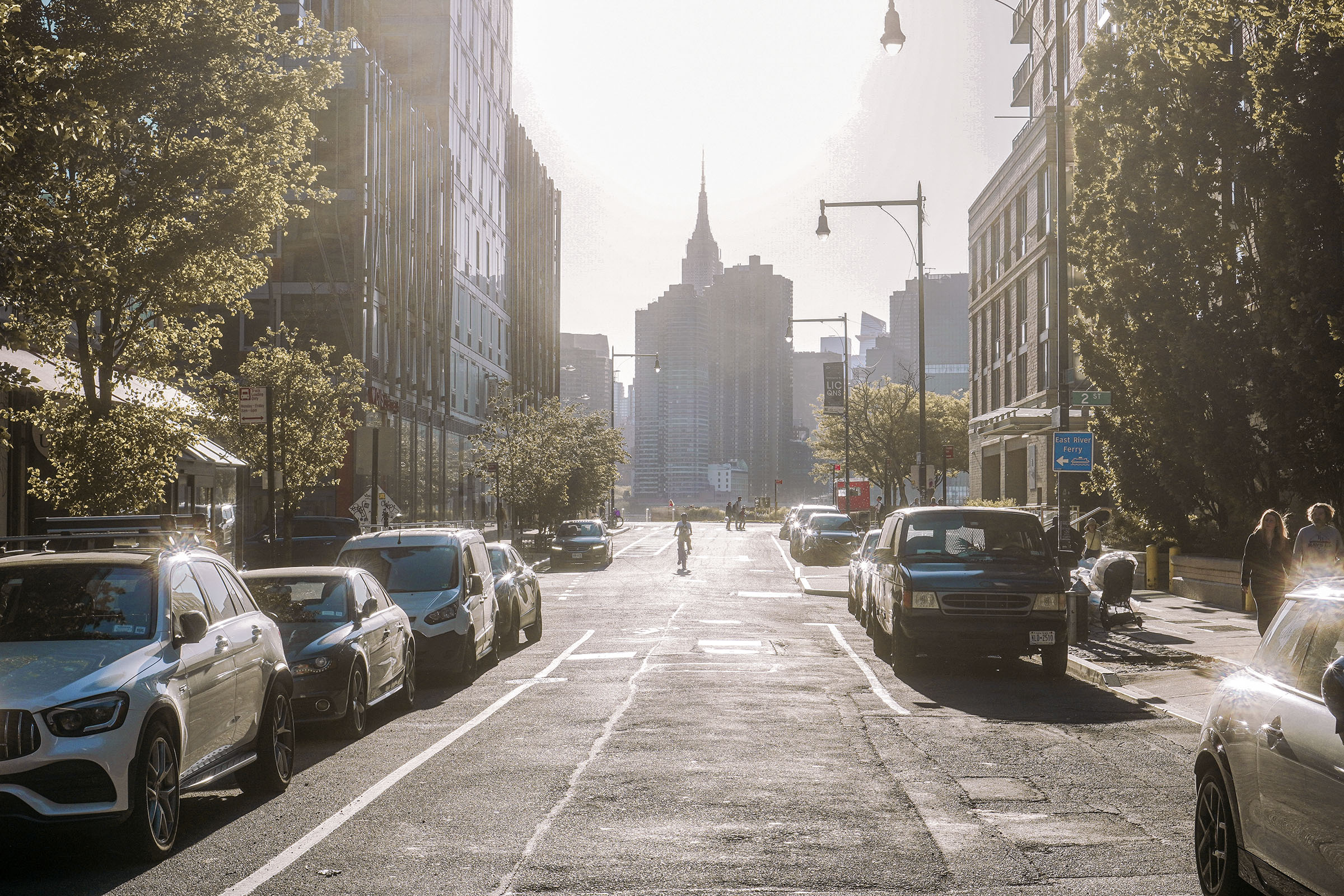 A cyclist riding toward the Empire State Building through a sunlit Long Island City street