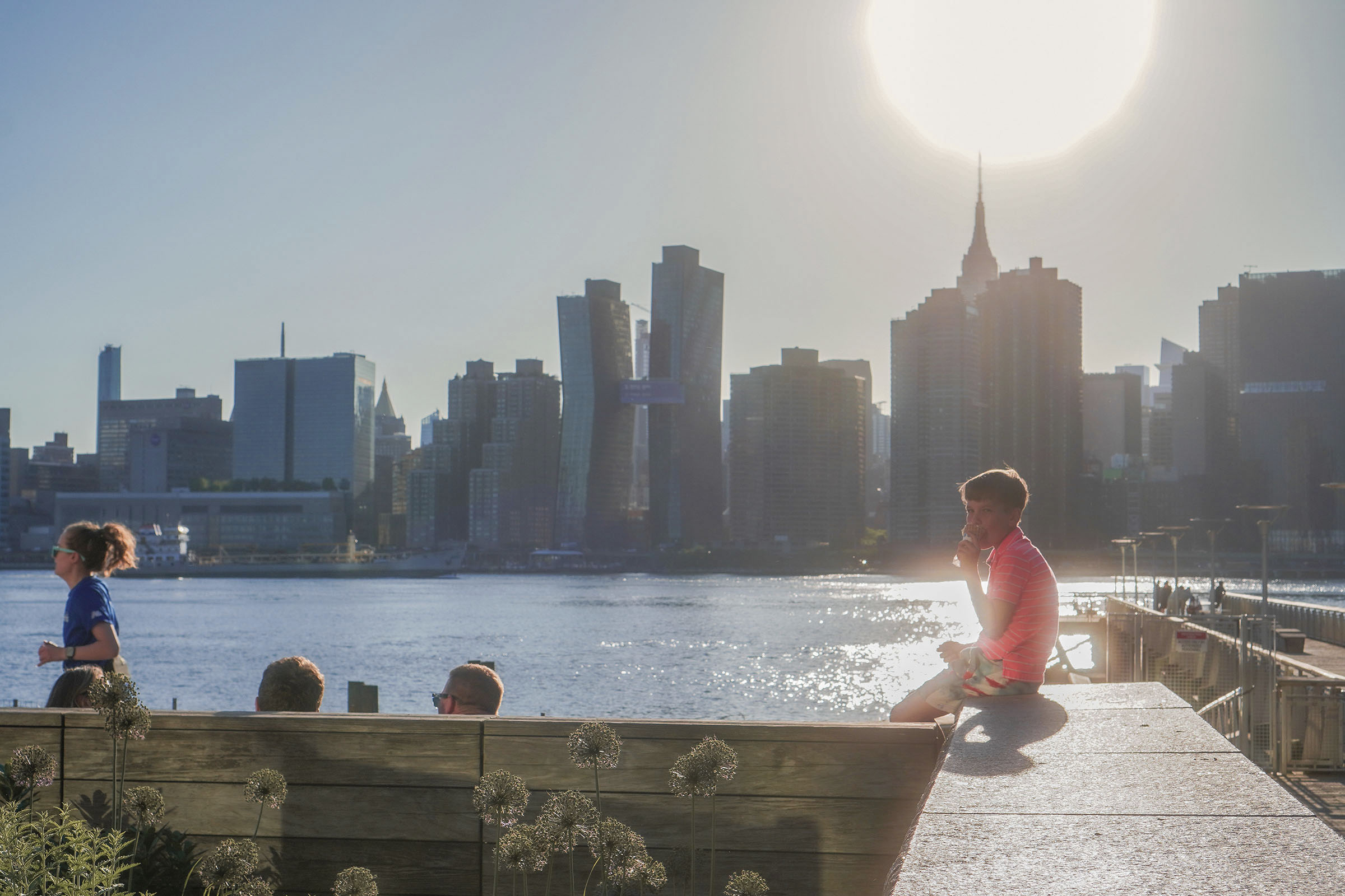 A sunlit New York skyline across the East River with people along the waterfront